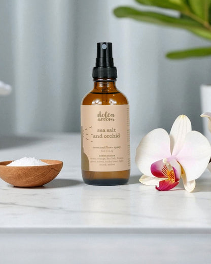 dolca aroom 4oz room and linen spray, sea salt and orchid scent on a marble surface with an orchid and wooden bowl.